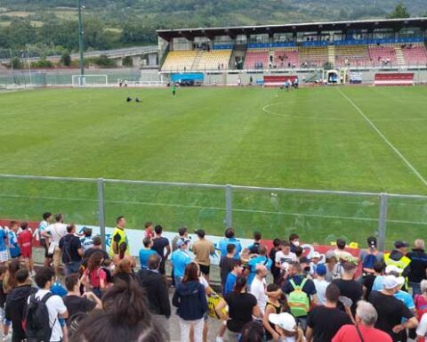 Tifosi del Napoli sugli spalti dello stadio Patini di Castel di Sangro durante un allenamento della squadra, con bandiere e sciarpe azzurre, in una giornata di sole