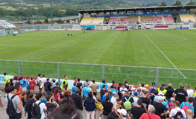 Tifosi del Napoli sugli spalti dello stadio Patini di Castel di Sangro durante un allenamento della squadra, con bandiere e sciarpe azzurre, in una giornata di sole