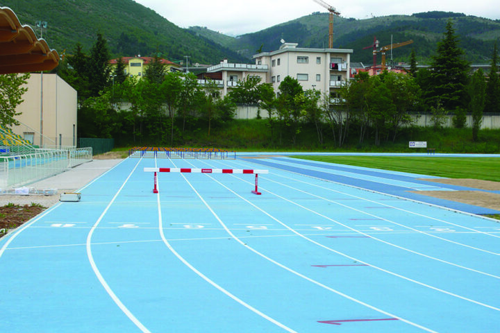 Vista sulle tribune e sulla pista di atletica color azzurro, immersa in uno scenario montano, con impianti e spalti recentemente rinnovati per ospitare allenamenti e competizioni.