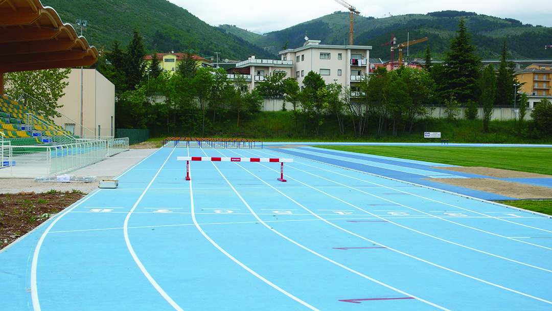 Vista sulle tribune e sulla pista di atletica color azzurro, immersa in uno scenario montano, con impianti e spalti recentemente rinnovati per ospitare allenamenti e competizioni.