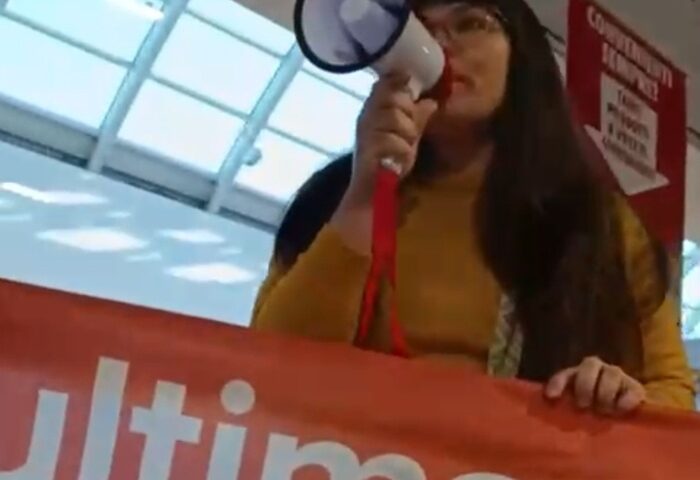 Un’attivista di “Ultima Generazione” con megafono e striscione arancione durante la protesta nel supermercato Conad dell’Aquila contro precarietà e salari bassi.