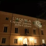 Facciata di Palazzo Margherita illuminata con la proiezione “L’Aquila Capitale italiana della Cultura”, simbolo delle iniziative in vista del 2026. Atmosfera serale durante uno degli eventi preparatori dedicati all’identità cittadina