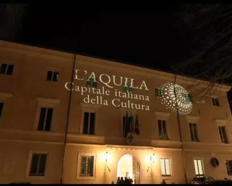 Facciata di Palazzo Margherita illuminata con la proiezione “L’Aquila Capitale italiana della Cultura”, simbolo delle iniziative in vista del 2026. Atmosfera serale durante uno degli eventi preparatori dedicati all’identità cittadina