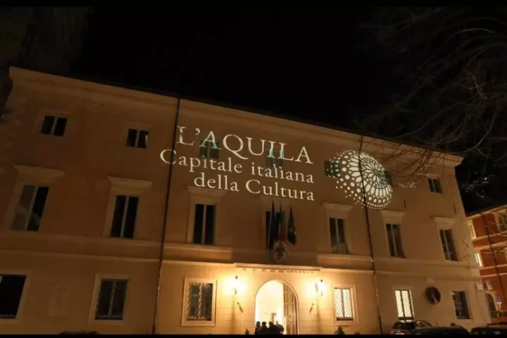 Facciata di Palazzo Margherita illuminata con la proiezione “L’Aquila Capitale italiana della Cultura”, simbolo delle iniziative in vista del 2026. Atmosfera serale durante uno degli eventi preparatori dedicati all’identità cittadina